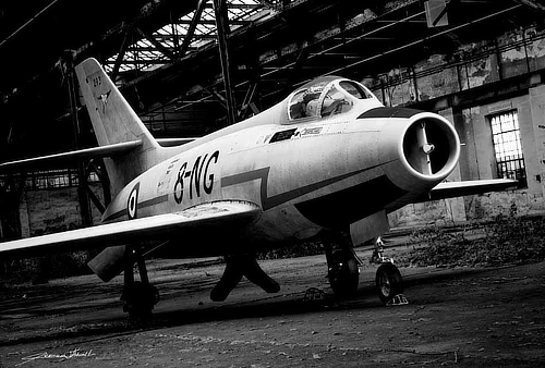 Mystère IV en clair-obscur, immobile dans un hangar abandonné. Une présence animale, la bête tapie dans sa tanière, révélée par la lumière.